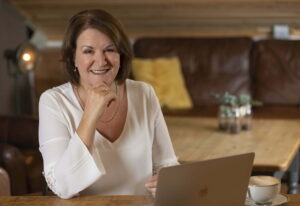Louise Jenner, The Dream Job Coach is seated at a table with her laptop open. She is wearing a white v neck blouse and is smiling into the camera, with her chin resting on her right hand.