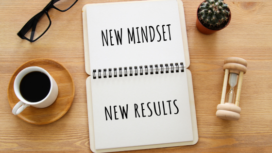 Wooden table top with specs, coffee, egg-timer and a note book with the words: NEW MINDSET NEW RESULTS